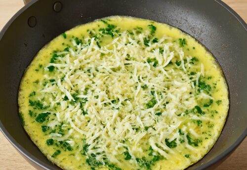 Quesadilla-2 Uncooked spinach and egg omelette in a non-stick pan, topped with shredded cheese, ready to cook for breakfast quesadilla.