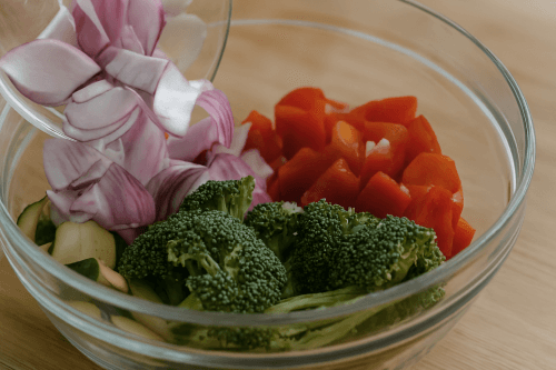 Chopped fresh vegetables on a light oak wood table, including half-moon zucchini slices, chunky red bell peppers, red onion wedges, and small broccoli florets, arranged for meal prep in a home kitchen setting.